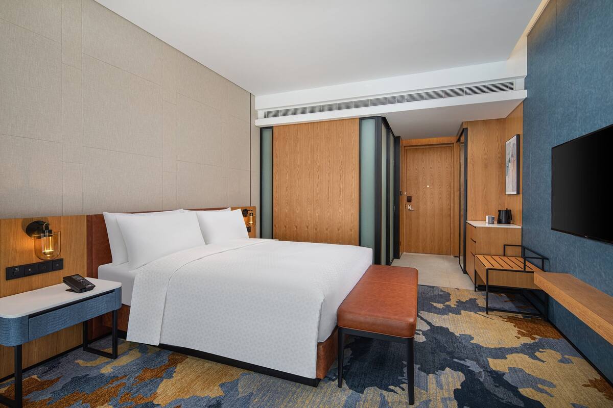 A hotel room with a bed, bedside table, bench, TV, and a small desk with a chair.