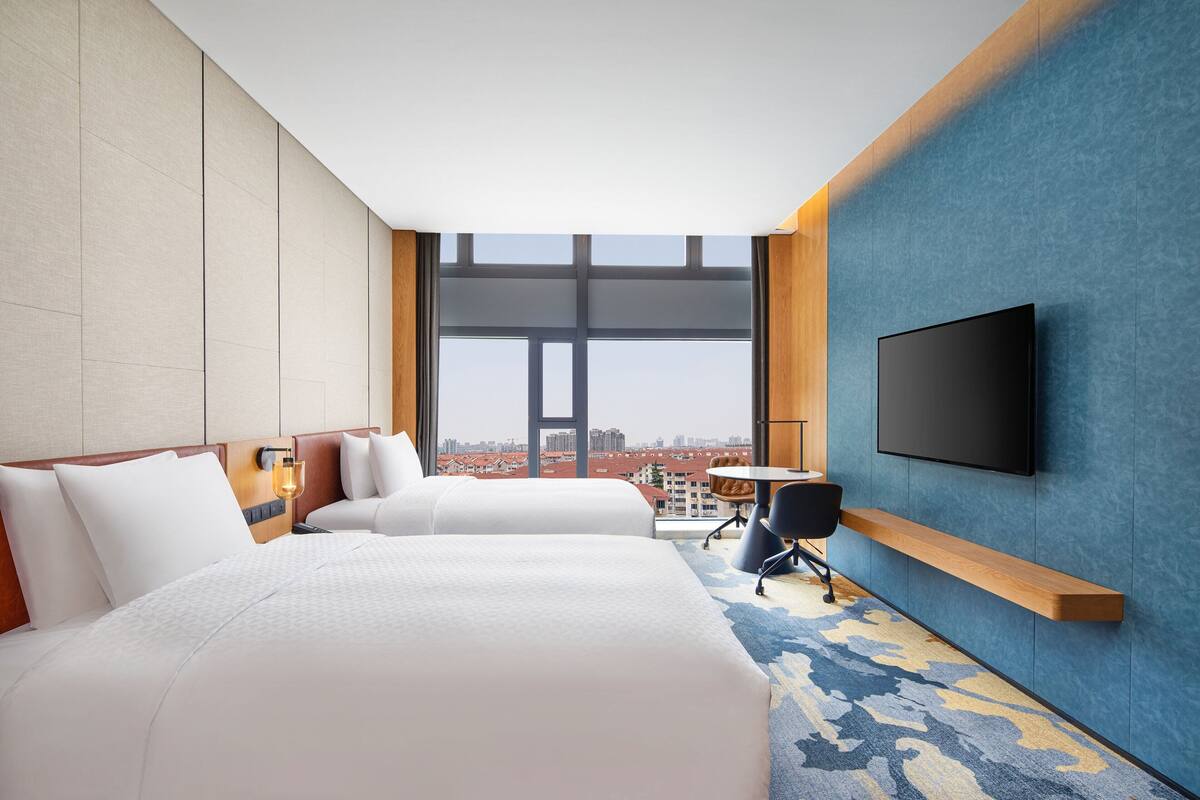 A hotel room with two beds, a TV, a desk, and a view of the city.