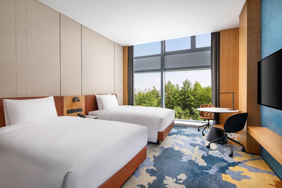 A hotel room with two beds, a desk, a chair, a TV, and a large window with a city view.