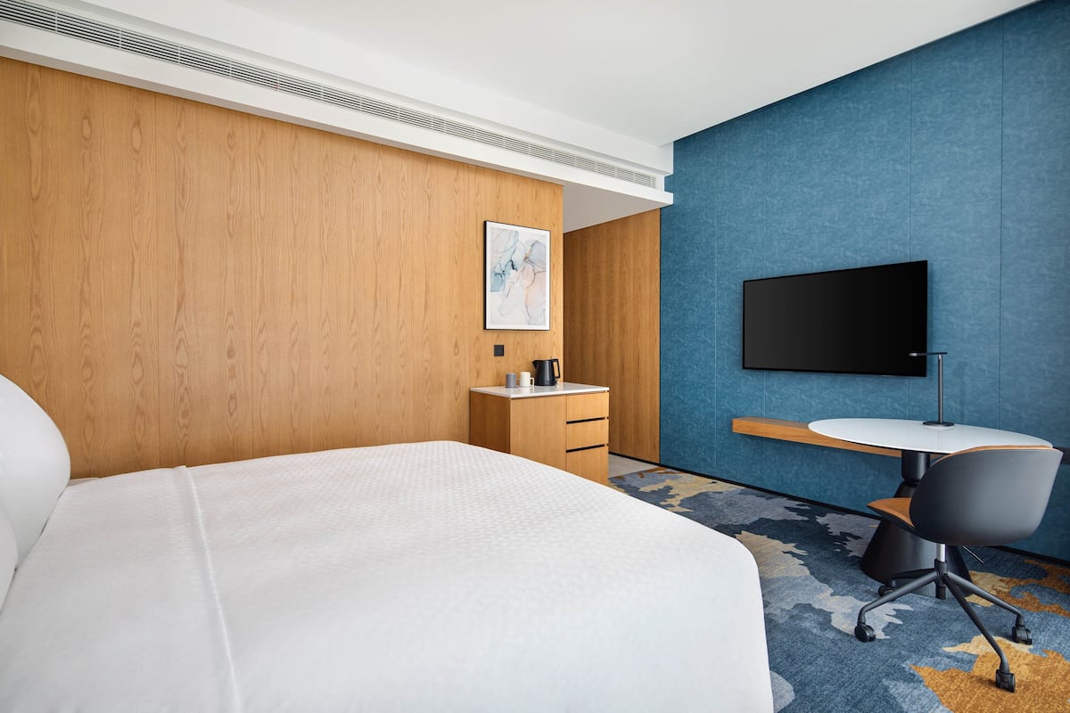 A hotel room with a large bed, a wooden nightstand, a desk with a chair, a flat-screen TV, and a blue accent wall.