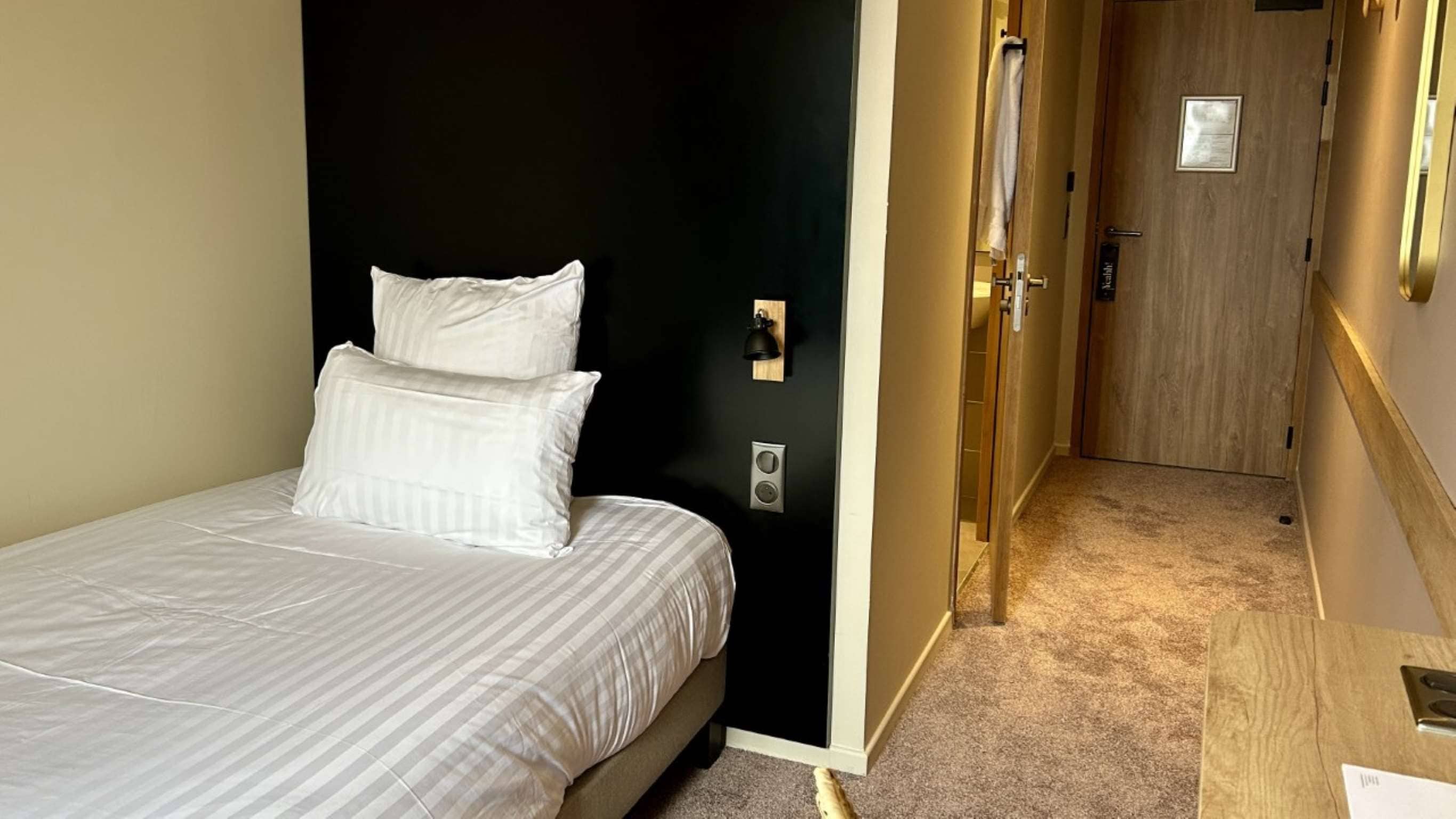 Ein Hotelzimmer mit einem Bett, einer Wandleuchte und einer Tür zu einem Flur.