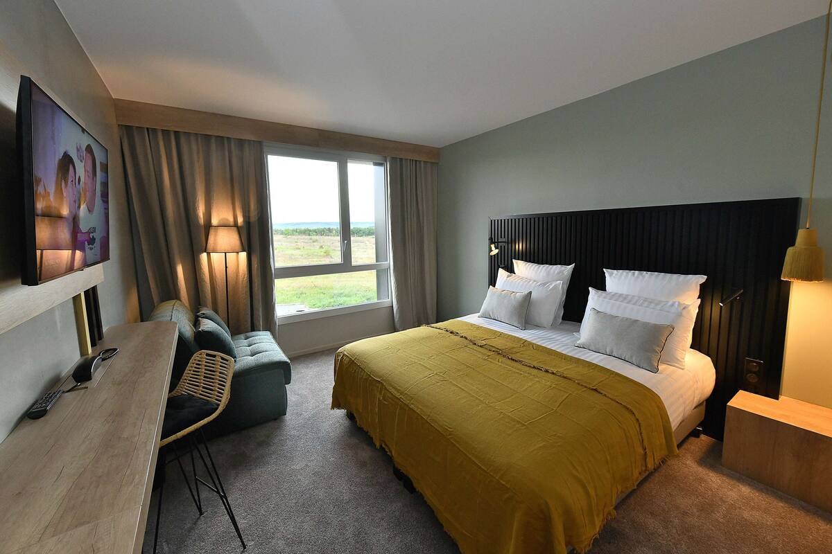 Une chambre d’hôtel avec un lit, un bureau et une chaise, une télévision et une fenêtre avec des rideaux.