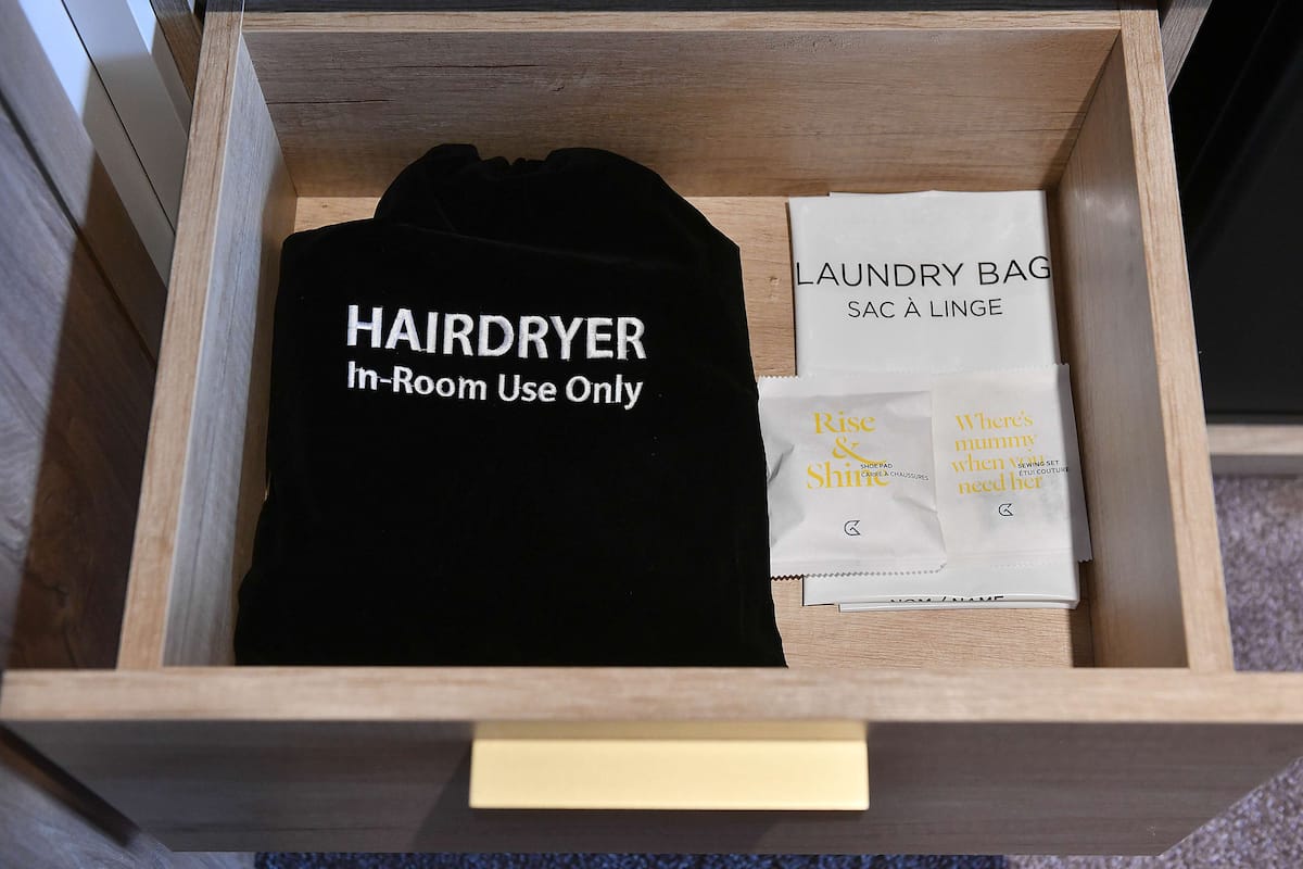 Un tiroir contenant un vêtement noir étiqueté « SÈCHE-CHEVEUX – Usage exclusif dans la chambre » et un sac à linge blanc avec du texte.