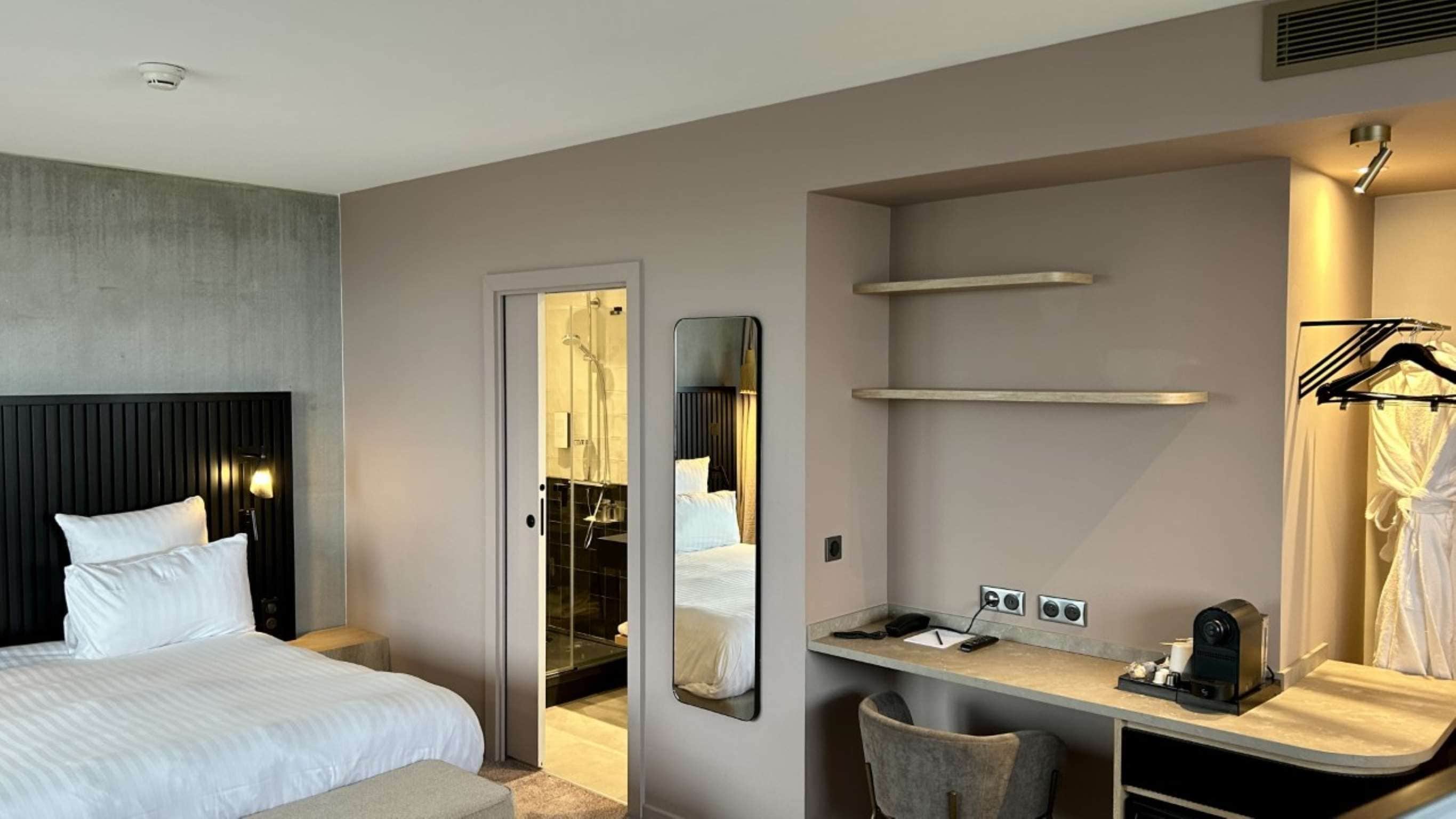 Habitación de hotel con cama, escritorio y silla, espejo, bata colgada y zona de ducha.