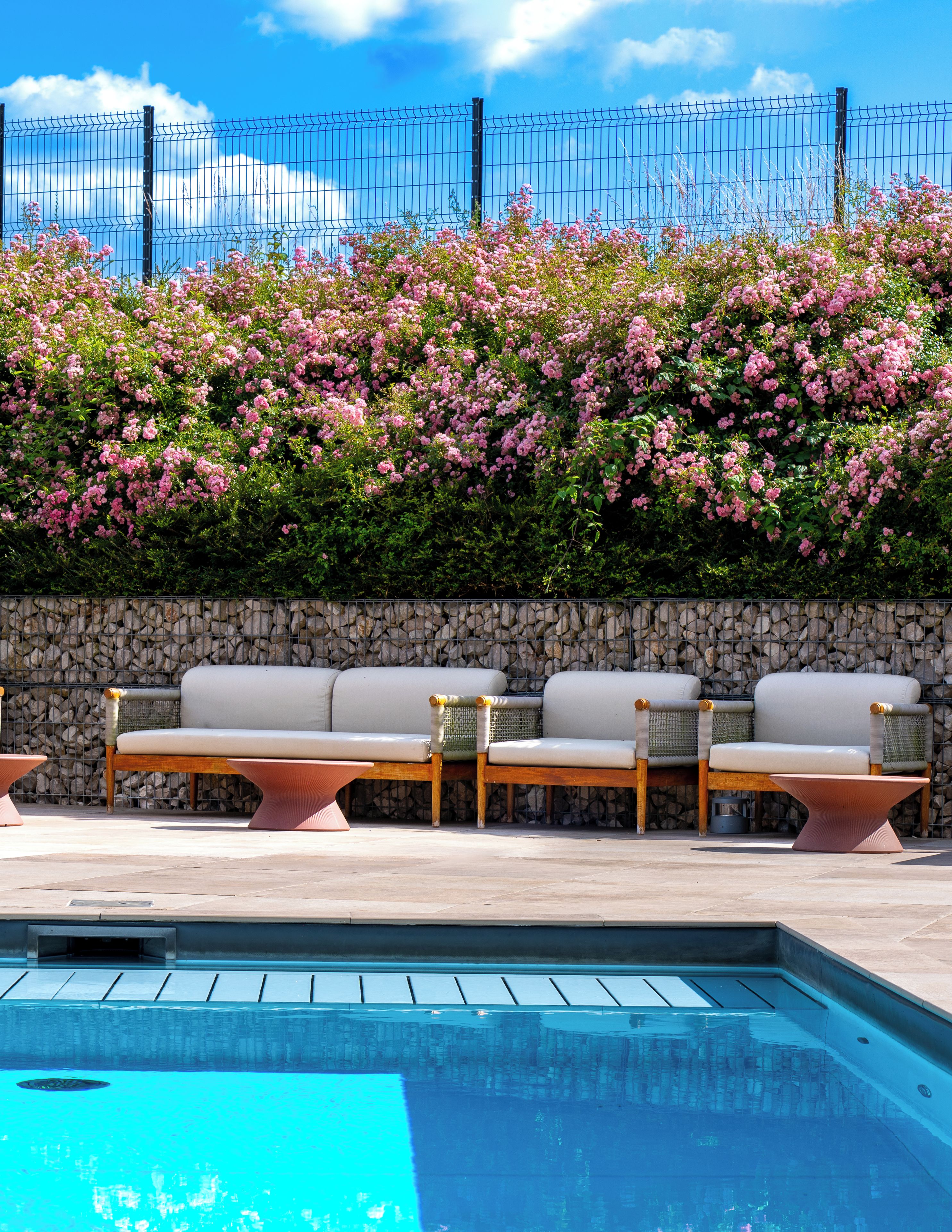 A pool area with a stone wall, a row of white cushioned chairs, and a metal fence with pink flowering bushes.