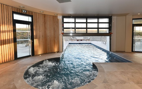 A modern indoor swimming pool with a view of the outdoors, wooden paneling on the walls, and a seating area visible through the windows.
