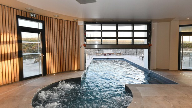 A modern indoor swimming pool with a view of the outdoors, wooden paneling on the walls, and a seating area visible through the windows. - Golden Tulip Reims (Bezannes)
