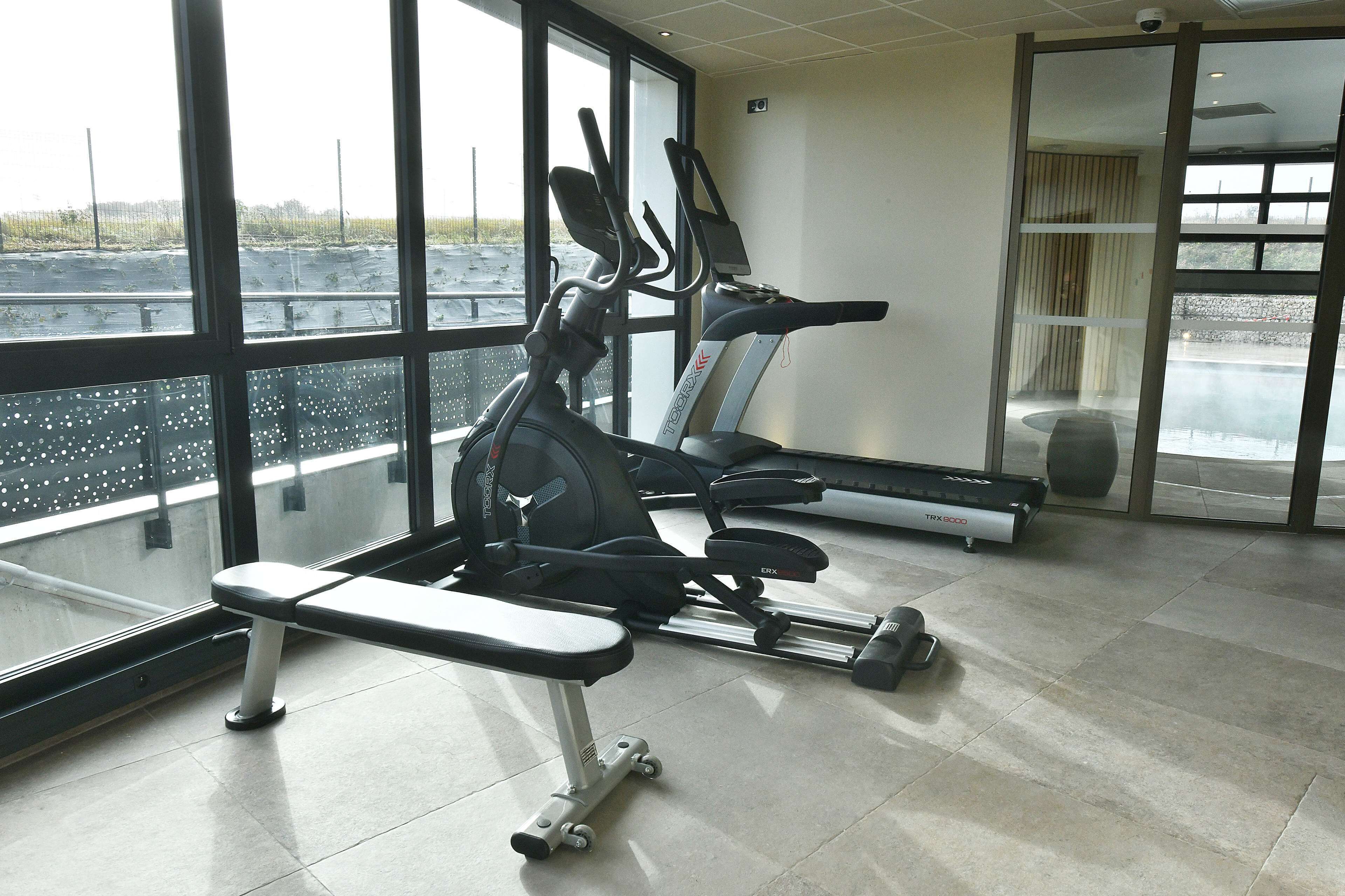 Een sportschool met fitnessapparatuur, grote ramen en uitzicht op een parkeerplaats.