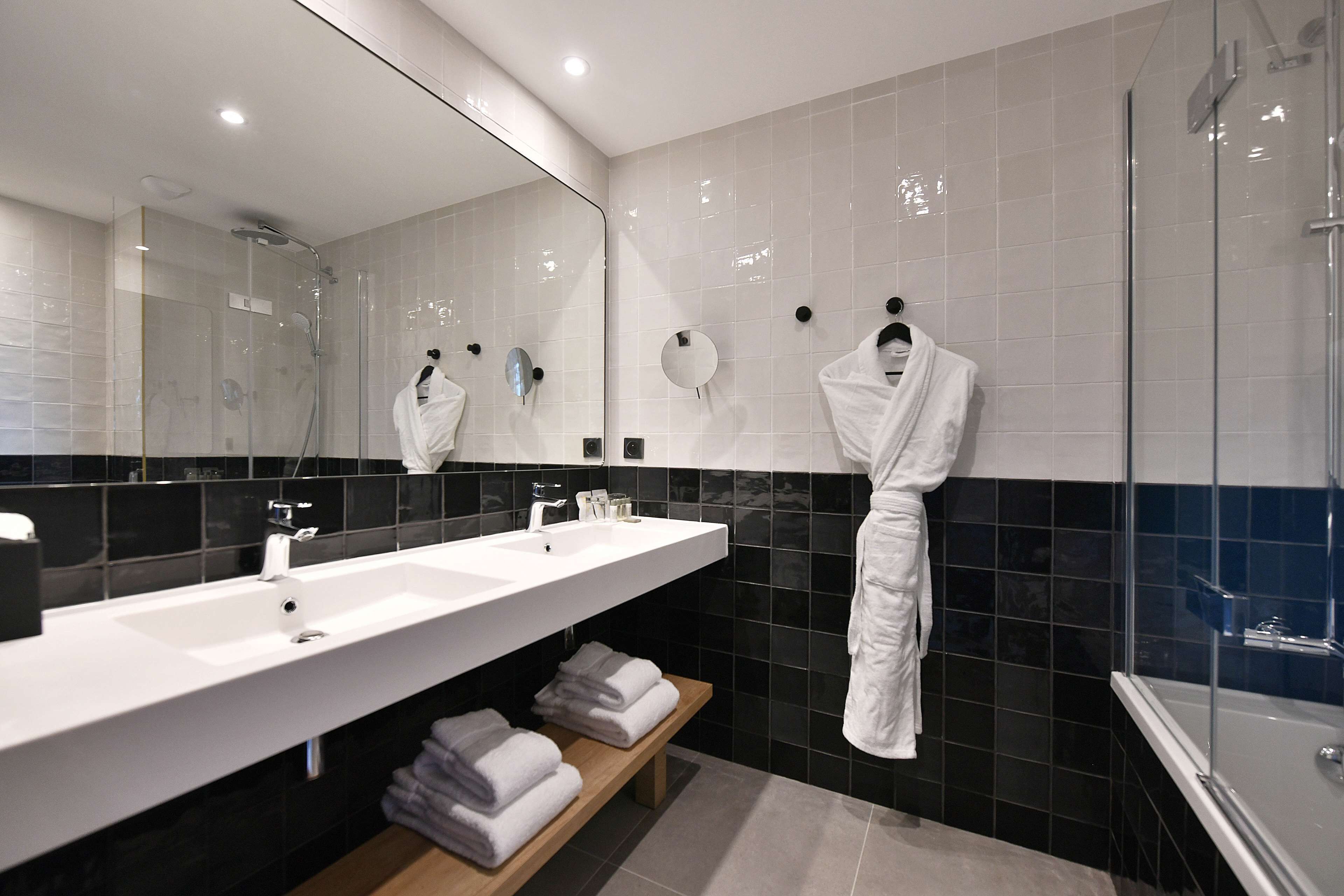 A modern bathroom with a large mirror, two sinks, a shower, and a bench with towels.