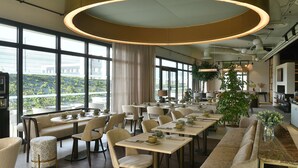 A spacious dining area with multiple tables and chairs, large windows offering a view of greenery outside, and modern pendant lighting. - Golden Tulip Reims (Bezannes)