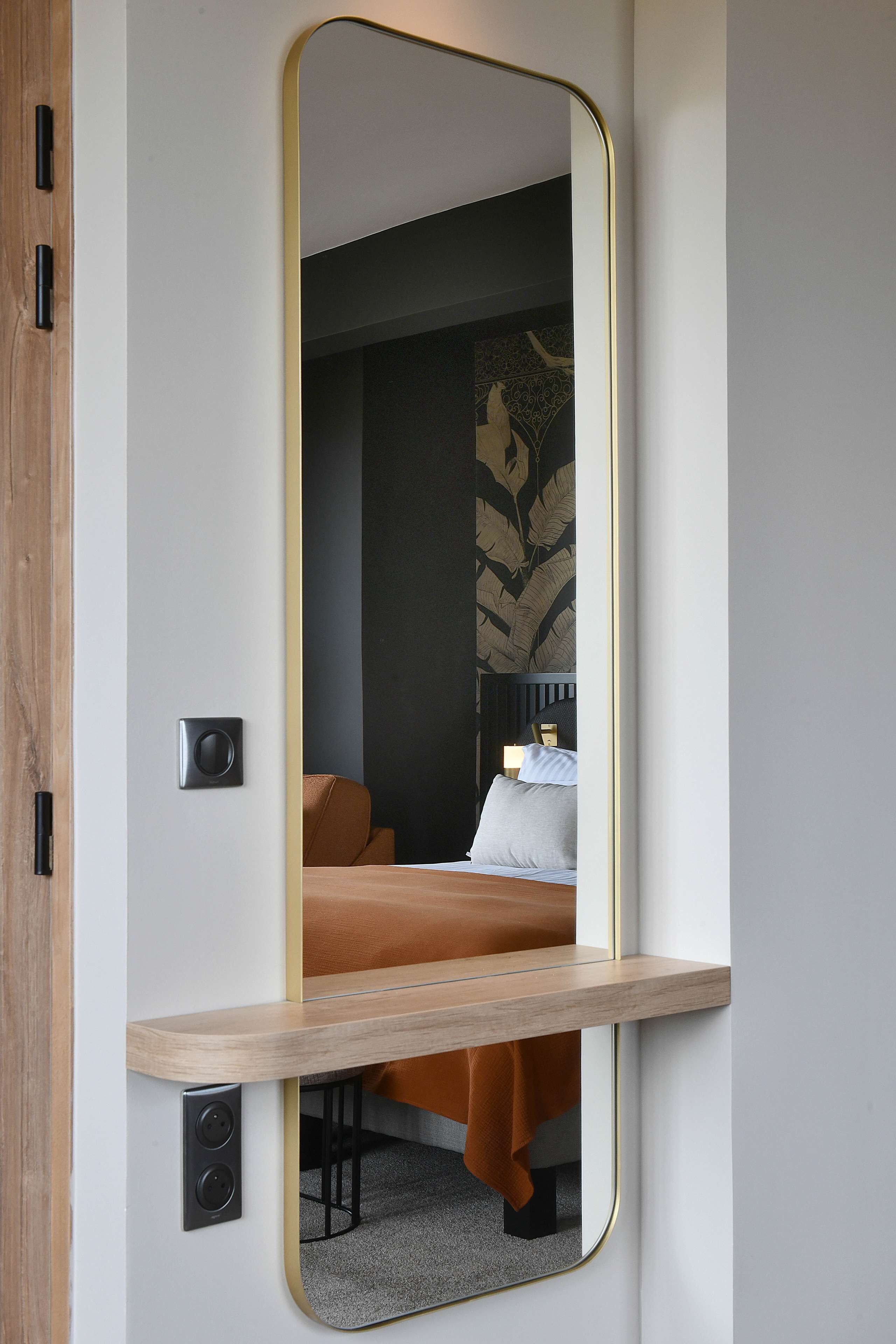 Una habitación de hotel moderna con un espejo grande, una mesita de noche de madera y una cama con manta marrón.
