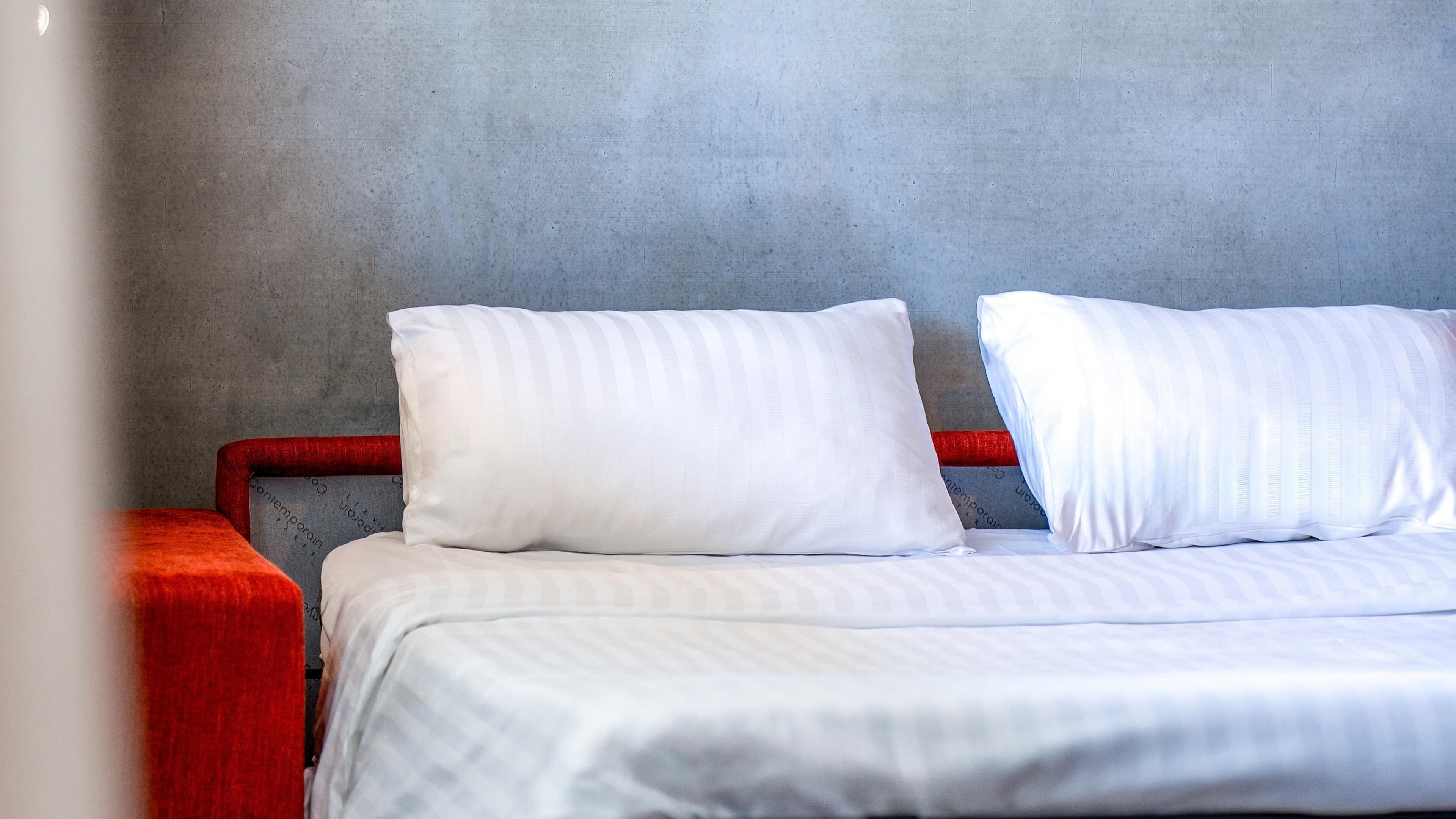 A bed with white bedding and two pillows, a red upholstered headboard, and a grey textured wall.