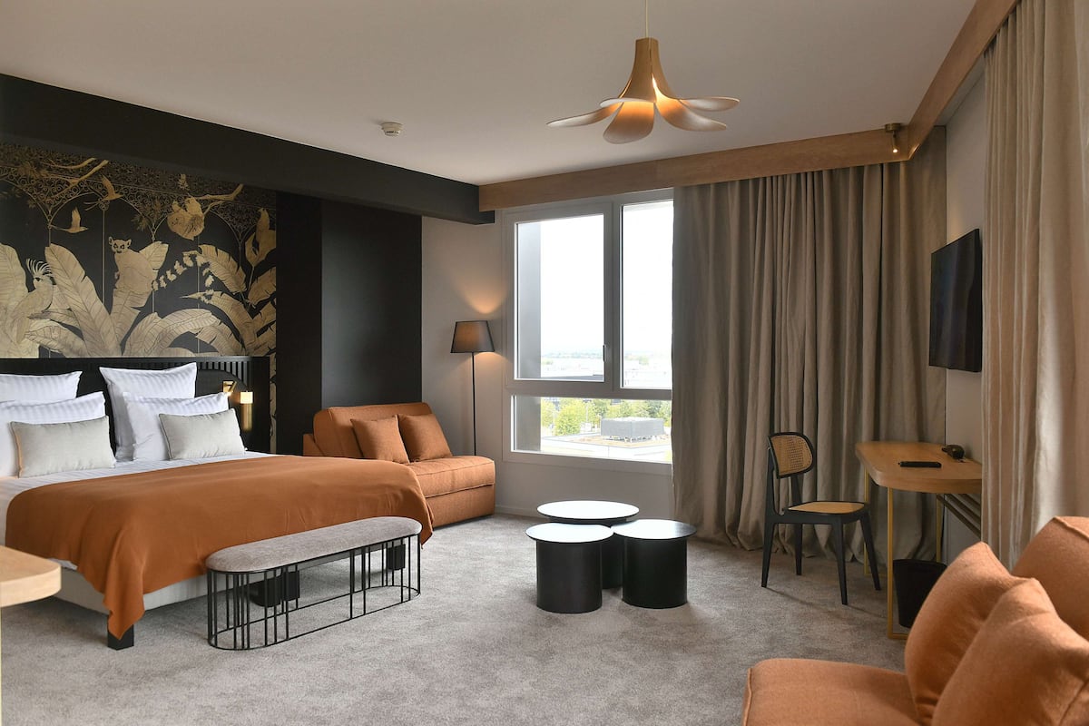 Une chambre d’hôtel moderne, dotée d’un grand lit, d’un canapé, d’un bureau et d’une chaise. On y trouve une fenêtre avec des rideaux, un ventilateur de plafond et un mur orné d’une fresque décorative.