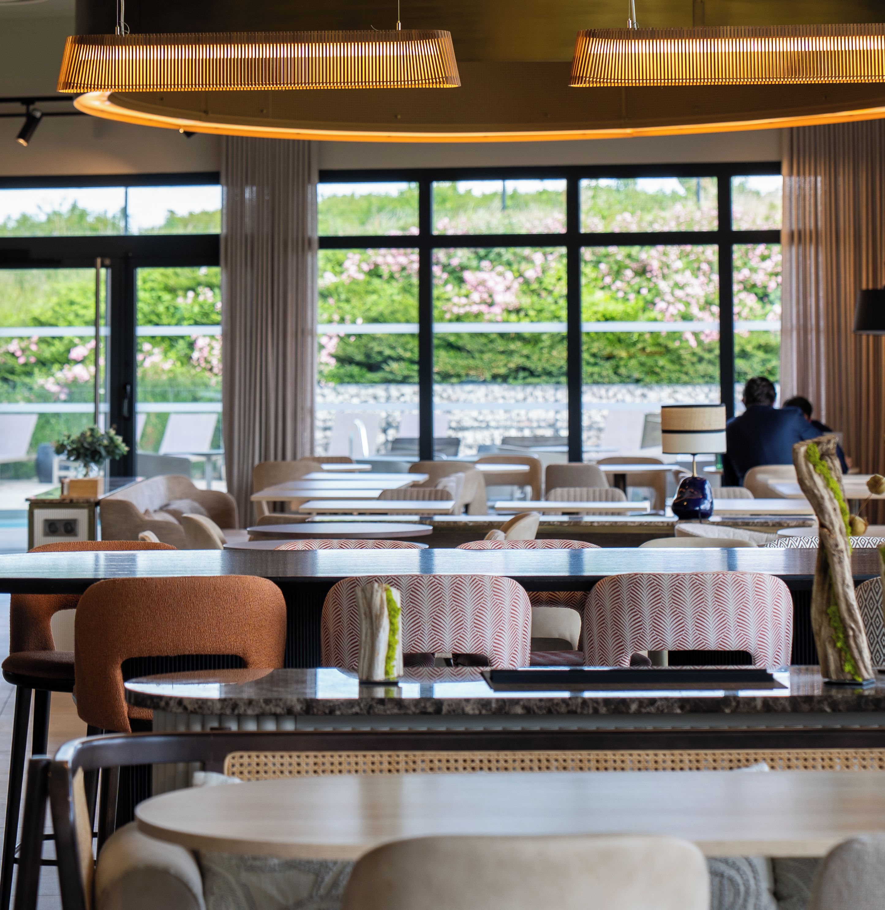 Un restaurante con una decoración moderna, que presenta una combinación de mesas redondas y rectangulares, y diversas opciones de asientos, incluidas cabinas y sillas. Amplios ventanales ofrecen vistas al exterior.