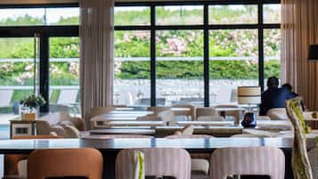 Un restaurante con una decoración moderna, que presenta una combinación de mesas redondas y rectangulares, y diversas opciones de asientos, incluidas cabinas y sillas. Amplios ventanales ofrecen vistas al exterior.