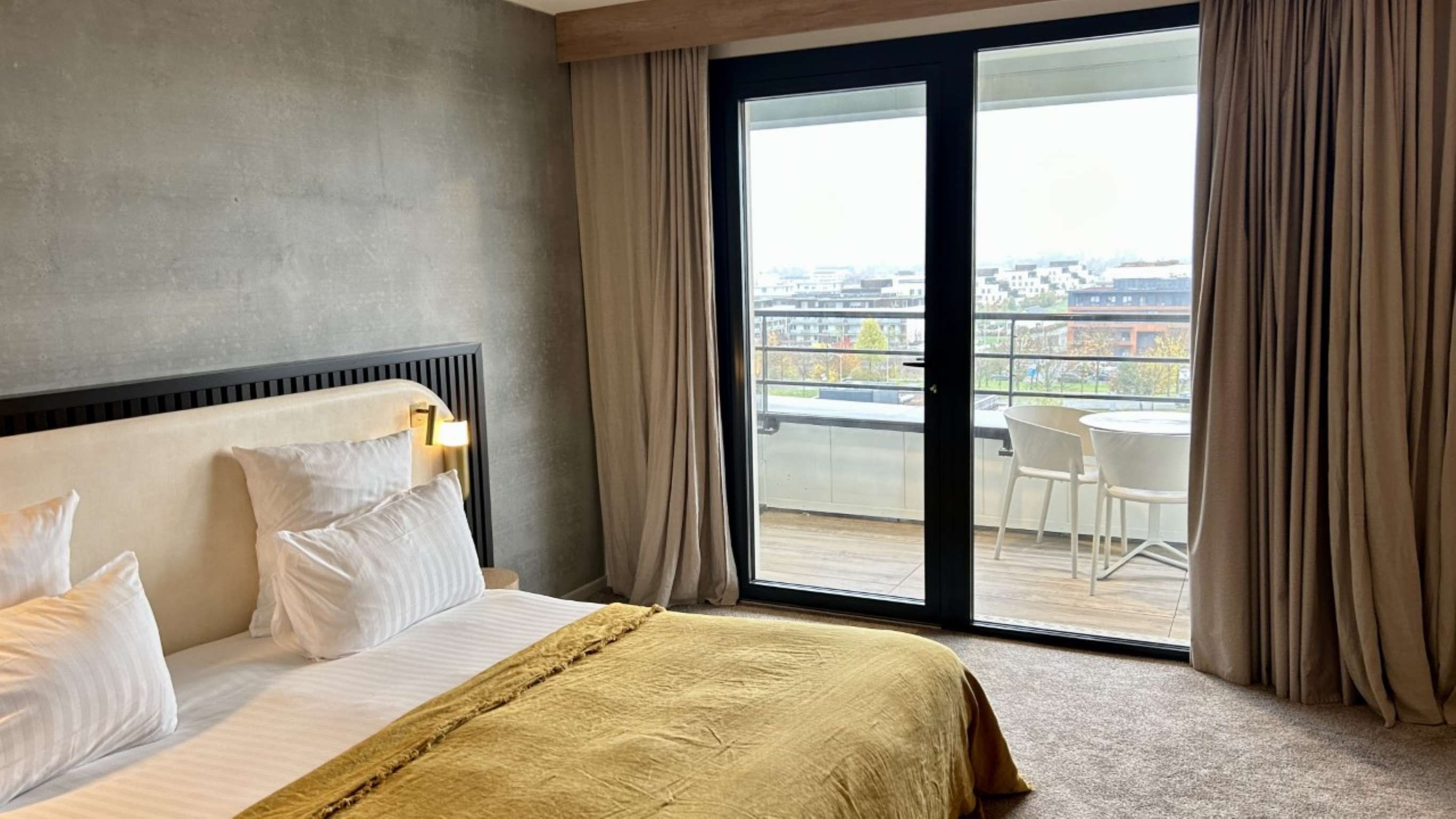 Een hotelkamer met een bed, uitzicht op de stad door een groot raam, en een balkon met stoelen.