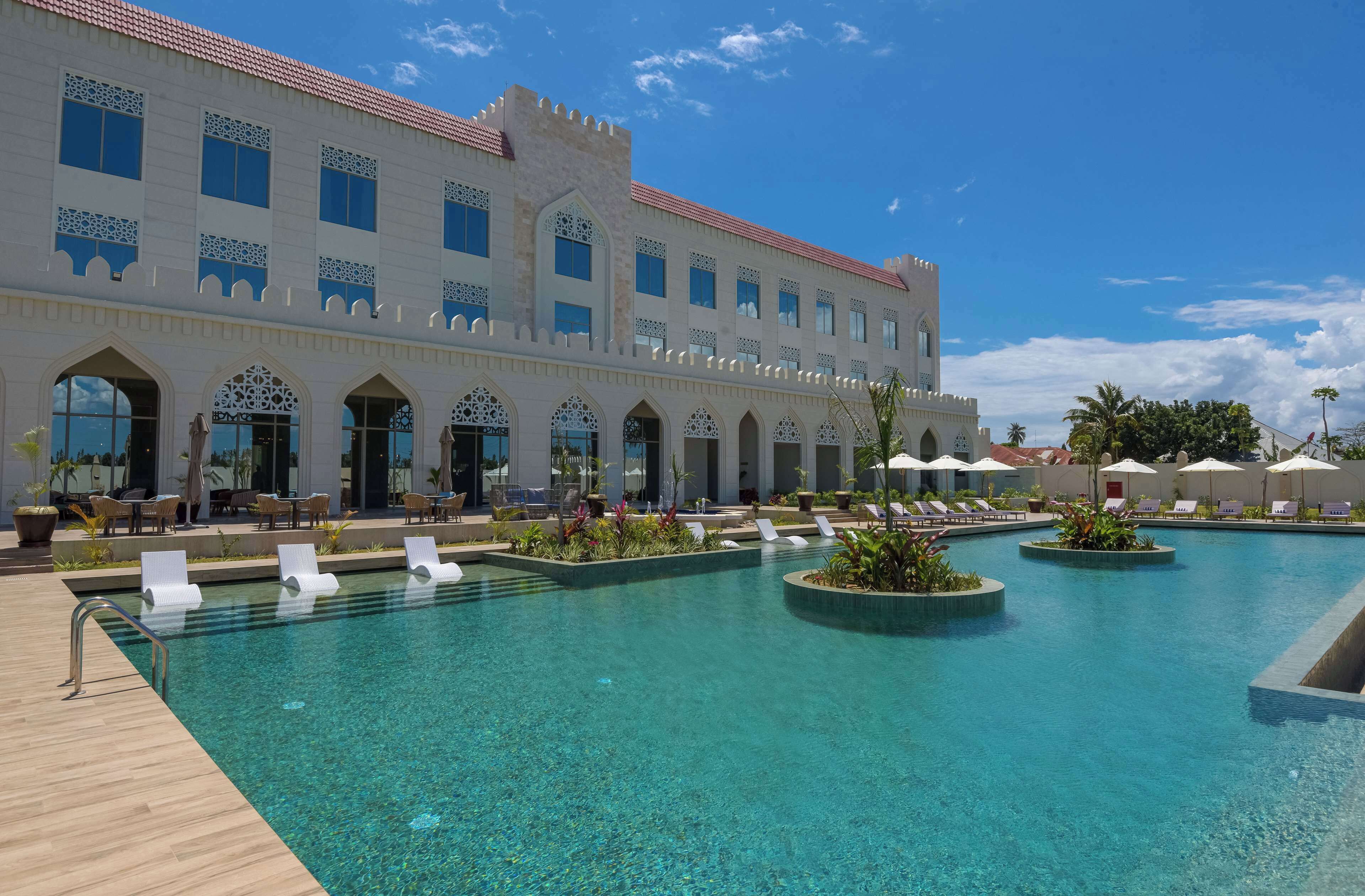 A swimming pool area with clear water, lounge chairs, and a large building with arched windows and a tiled roof.