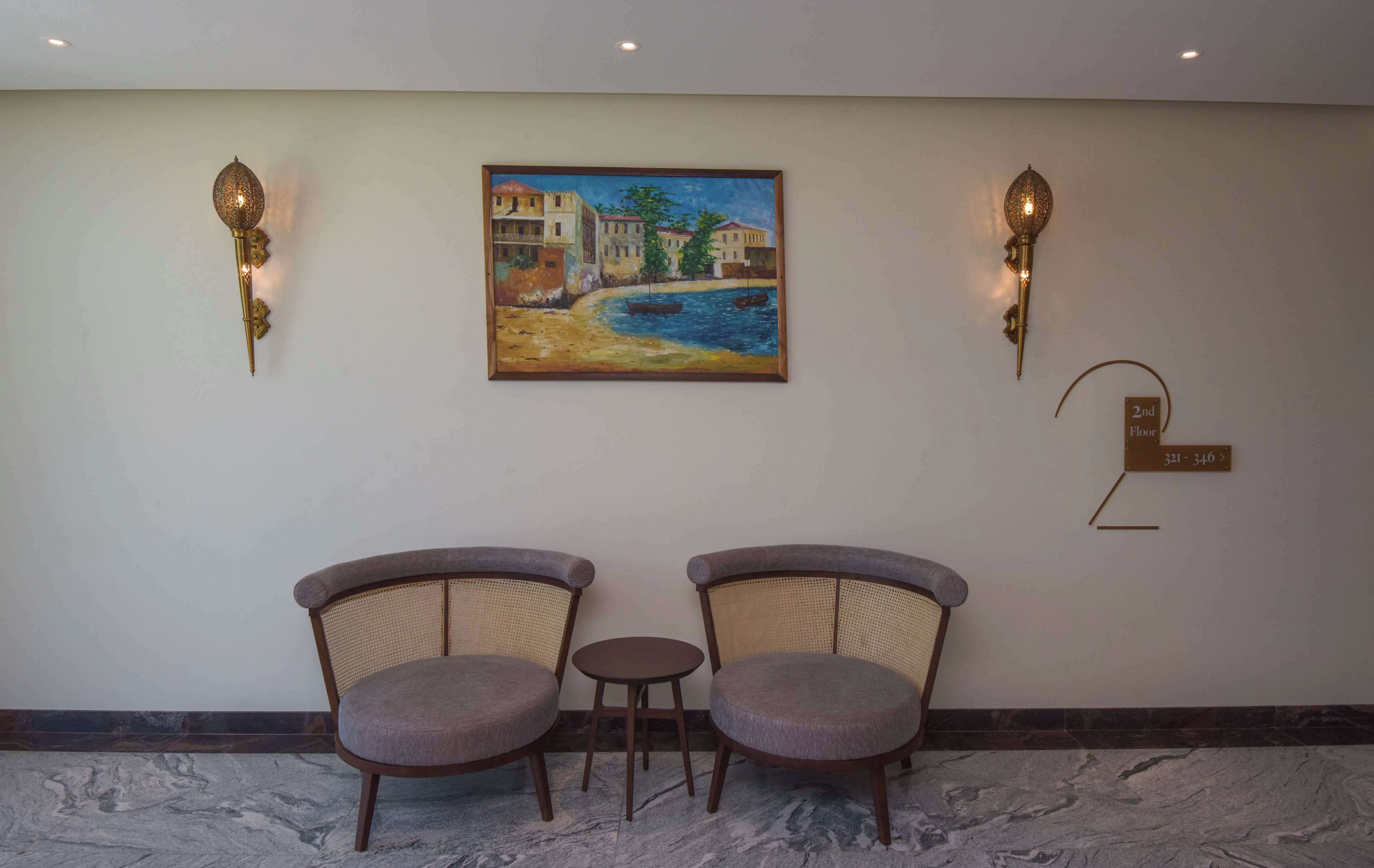 Un hall d’entrée avec deux fauteuils, une petite table, un tableau représentant un paysage de bord de mer, des appliques murales et un panneau indiquant le deuxième étage.