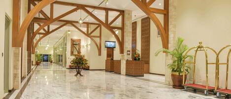 A spacious lobby with marble flooring, wooden beams, a reception desk, potted plants, and a television.