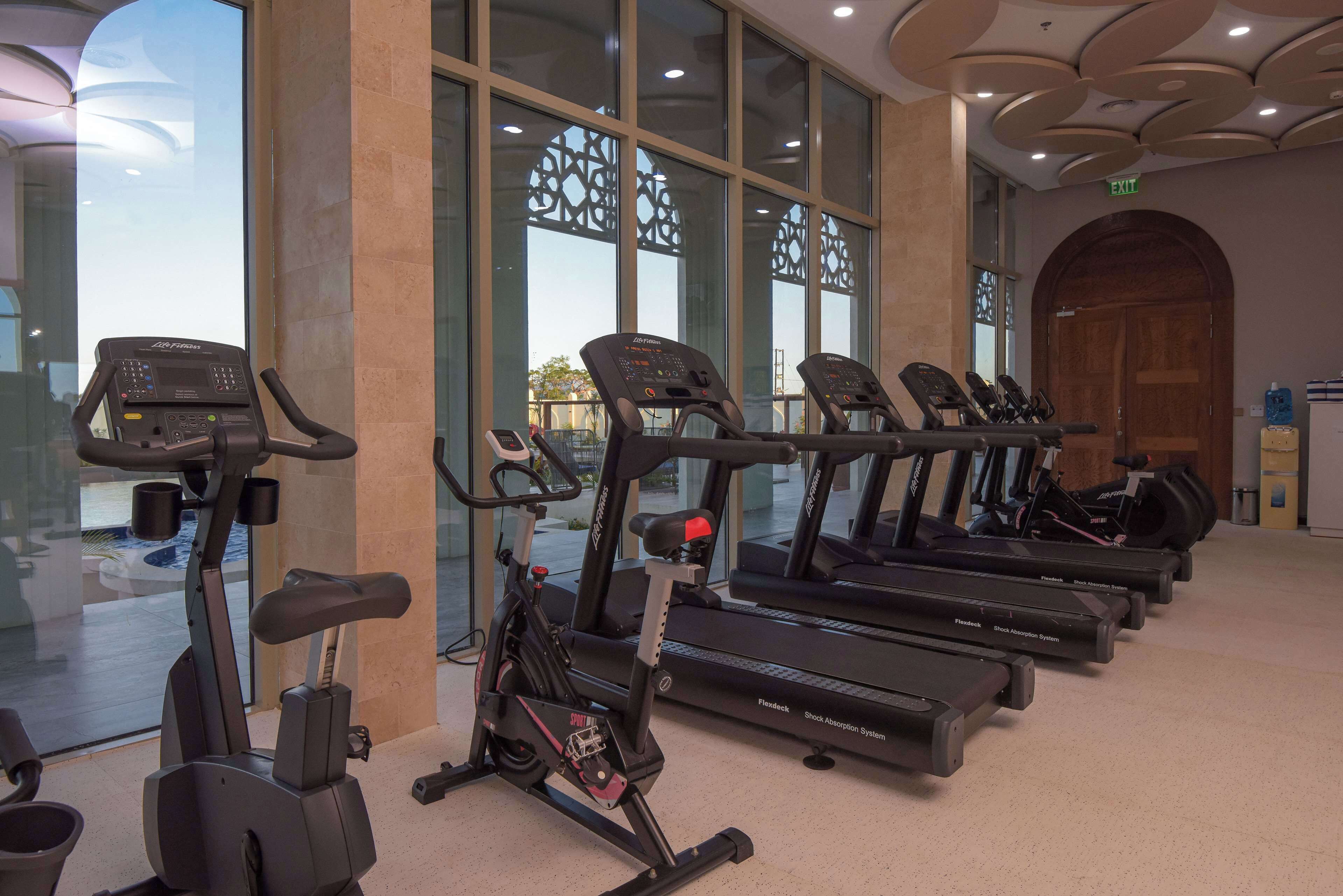 A row of stationary bikes and treadmills in a well-lit indoor area with large windows and a modern ceiling design.