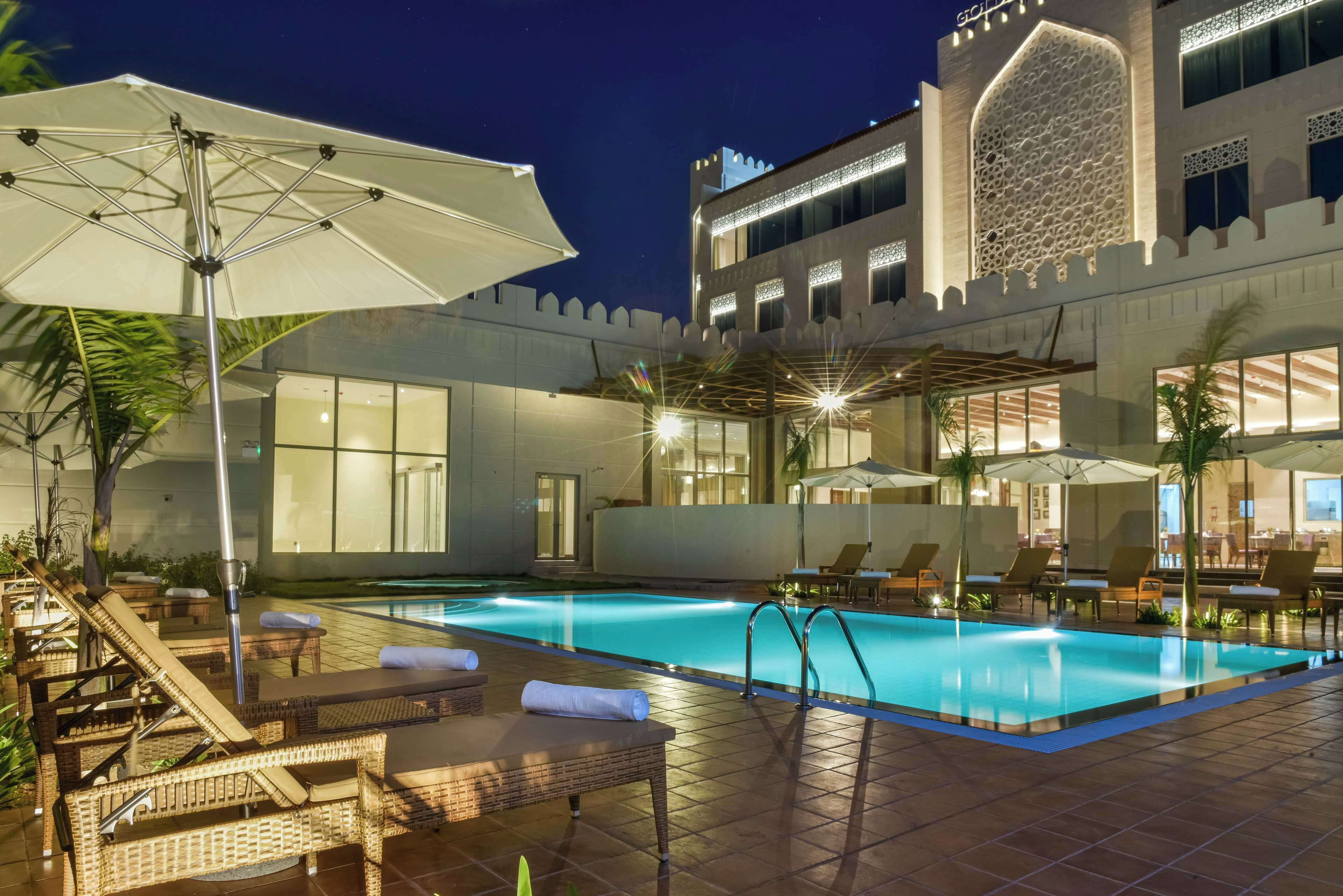 Zona de piscina con sillas de descanso y sombrillas, un edificio con fachada decorativa y cielo nocturno despejado.
