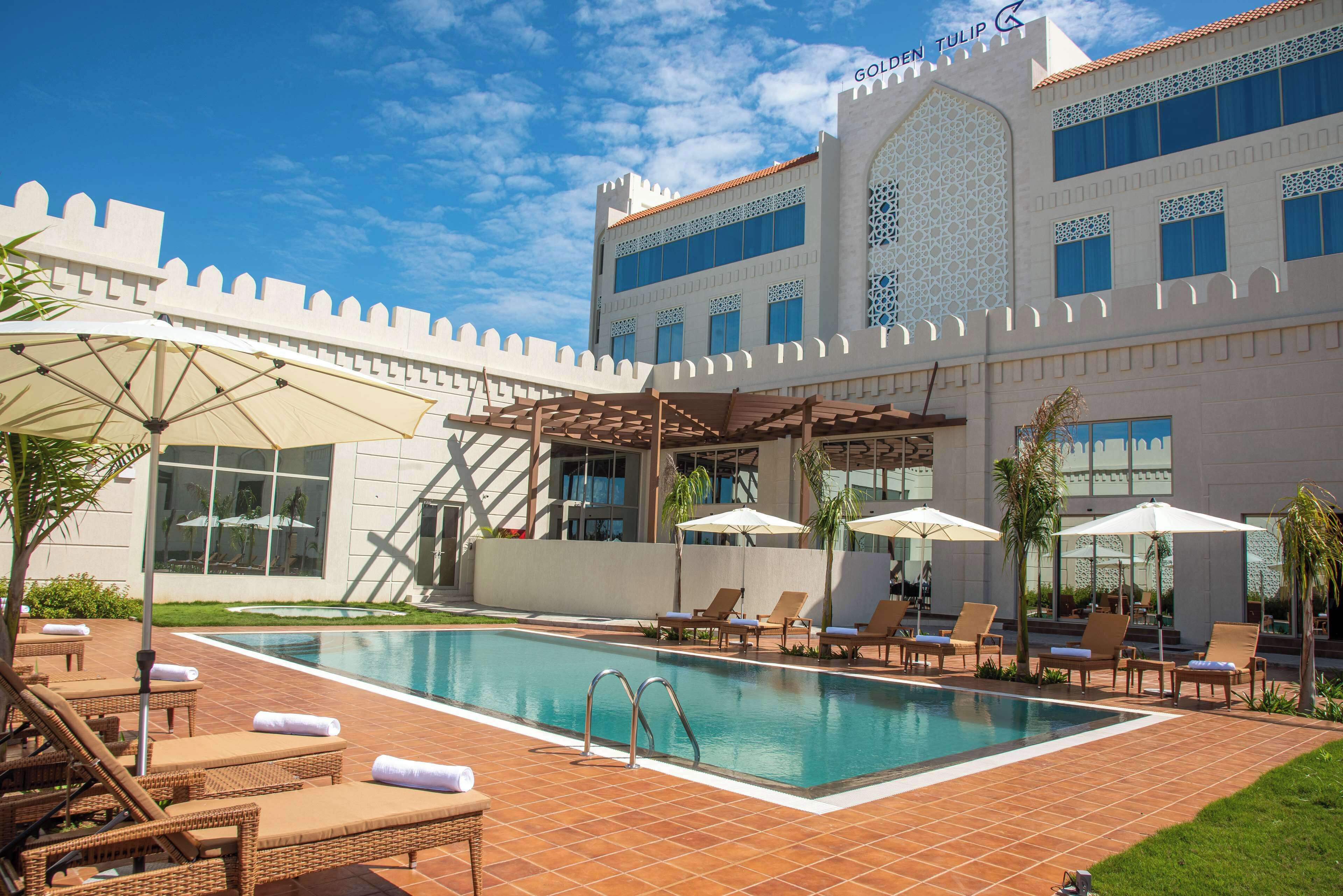 A pool area with lounge chairs and umbrellas, a clear sky, and a building with a castle-like facade.