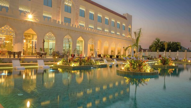 Un hotel con una piscina grande, varias zonas de descanso y una entrada imponente con ventanales arqueados.