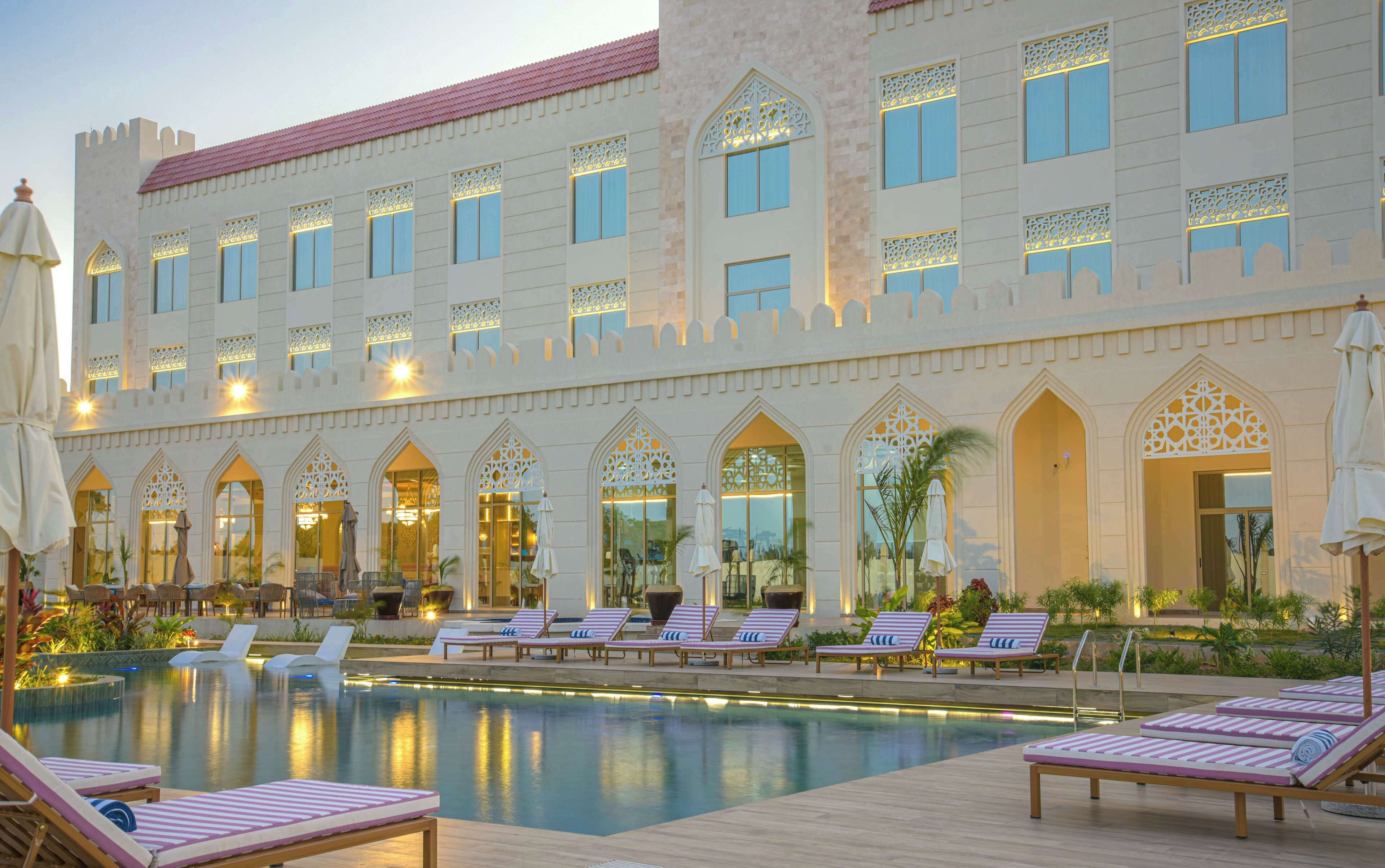A hotel with a pool area, lounge chairs, and a large building with arched windows and decorative lighting.