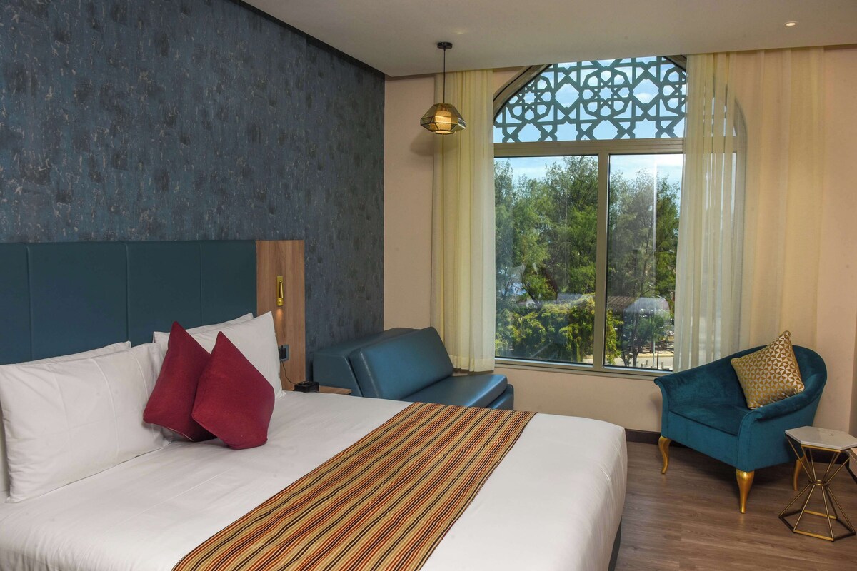 A hotel room with a large bed, a blue armchair, a small table, and a window with a view of trees.