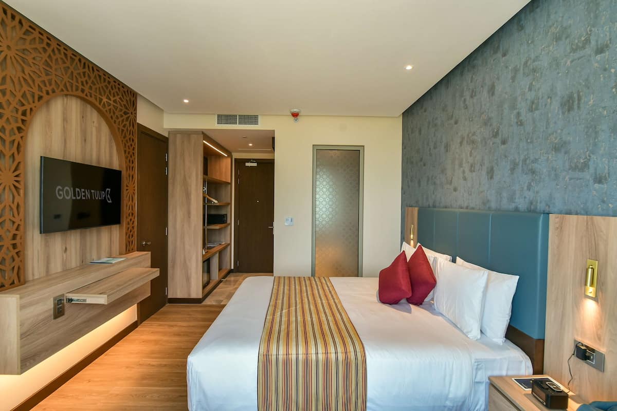 A modern hotel room with a large bed, a TV mounted on a wooden panel, a wardrobe, and a blue accent wall.