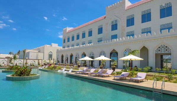A poolside area with lounge chairs and umbrellas, a large pool, and a building with arched windows and intricate designs. - Golden Tulip Zanzibar Airport (Zanzibar Town)