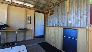 A wooden cabin with a metal-framed ceiling, a small table with a chair, a refrigerator, and a door. - Bunky on the Beach · UNESCO Geopark (Saint Martins Parish)