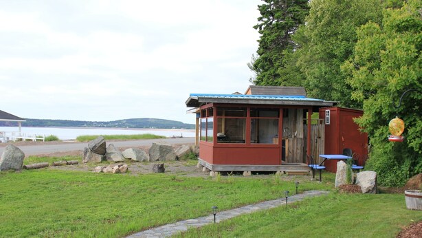 A small wooden cabin with a blue roof, situated on a grassy area near a body of water. - Bunky on the Beach · UNESCO Geopark (Saint Martins Parish)
