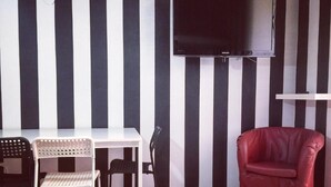 Huoneessa on mustavalkoraidallinen seinä, valkoinen pöytä, jonka yläpuolelle on asennettu litteänäyttöinen televisio, sekä punainen tuoli.