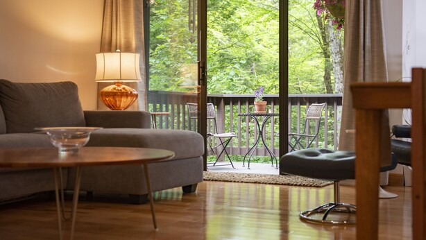 A cozy living room with a sofa, a coffee table, and a lamp. There is a balcony with a view of trees and a railing. - Vermont get-away end unit townhouse nestled in the trees  (Hartford)