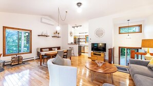 A spacious living room with a wooden floor, a dining area with a large window, and a kitchen in the background. - Vermont get-away end unit townhouse nestled in the trees  (Hartford)