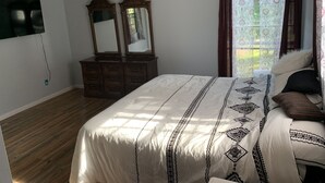A bedroom with a bed, a ceiling fan, a dresser, two mirrors, and a window with patterned curtains.