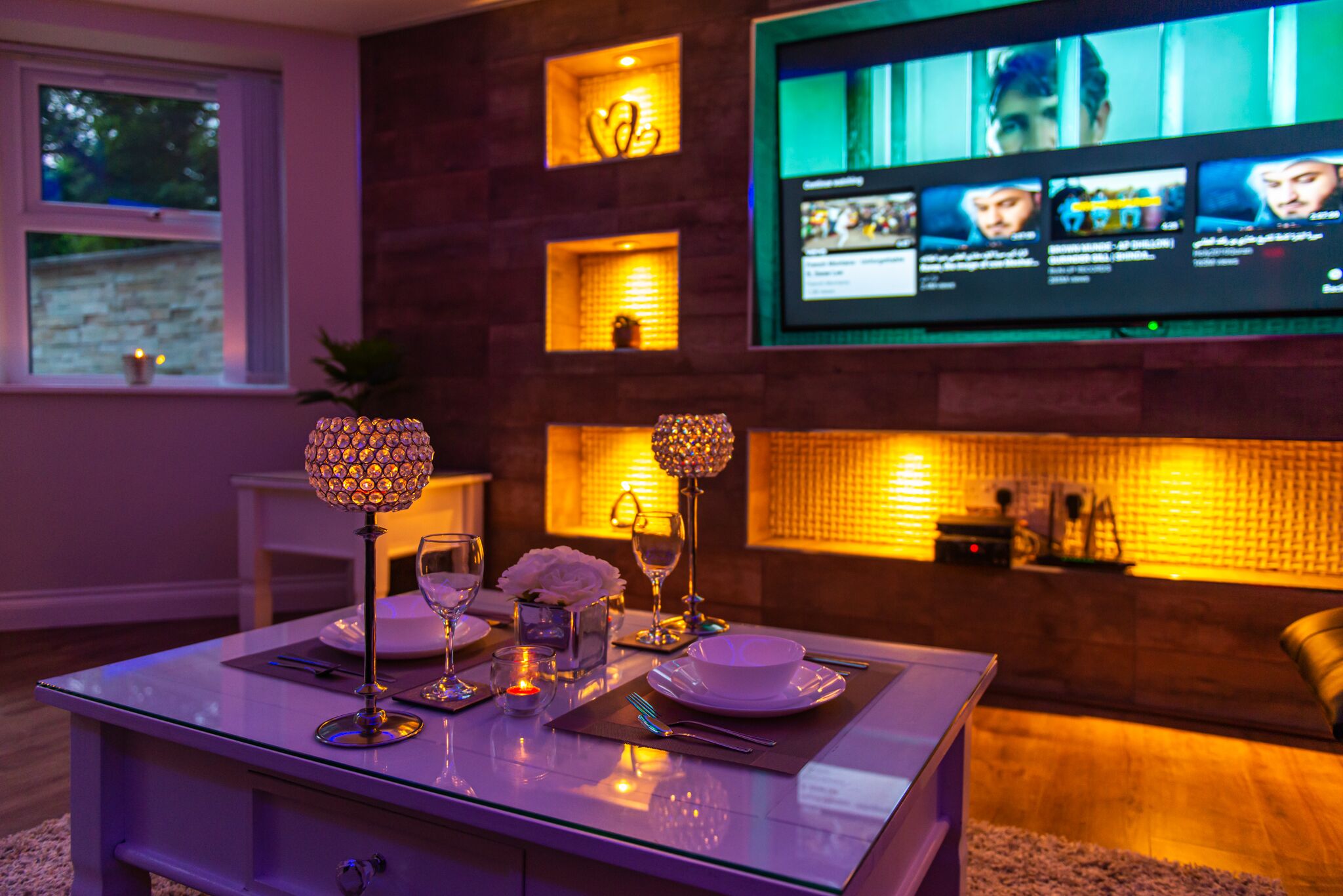 A modern living room with a glass coffee table, two decorative lamps, a flat-screen TV mounted on the wall, and a cozy seating area with a cushioned bench.