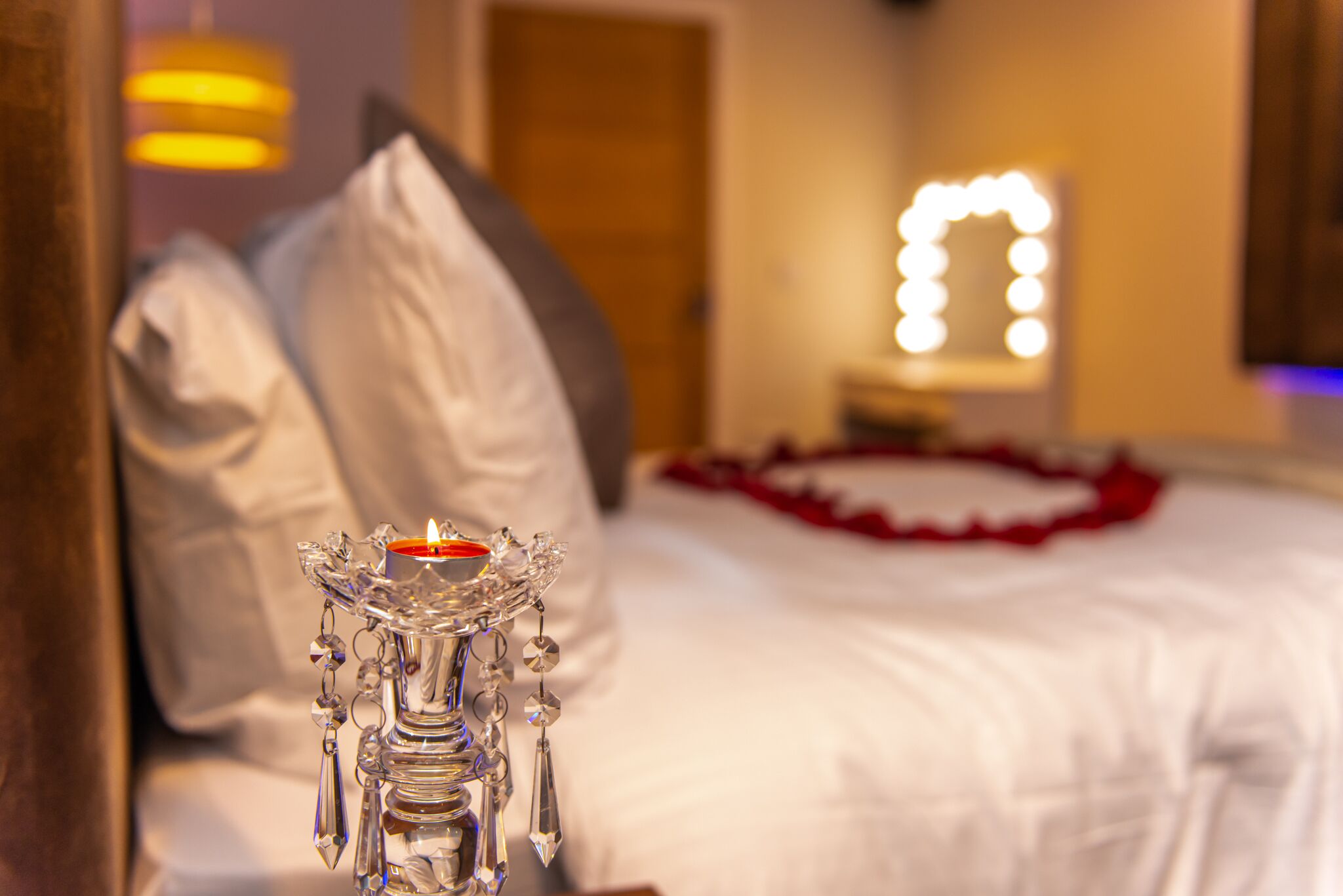 Una cama con ropa de cama blanca y un borde decorativo rojo, una mesita de noche con un candelabro de cristal y un espejo con luces al fondo.