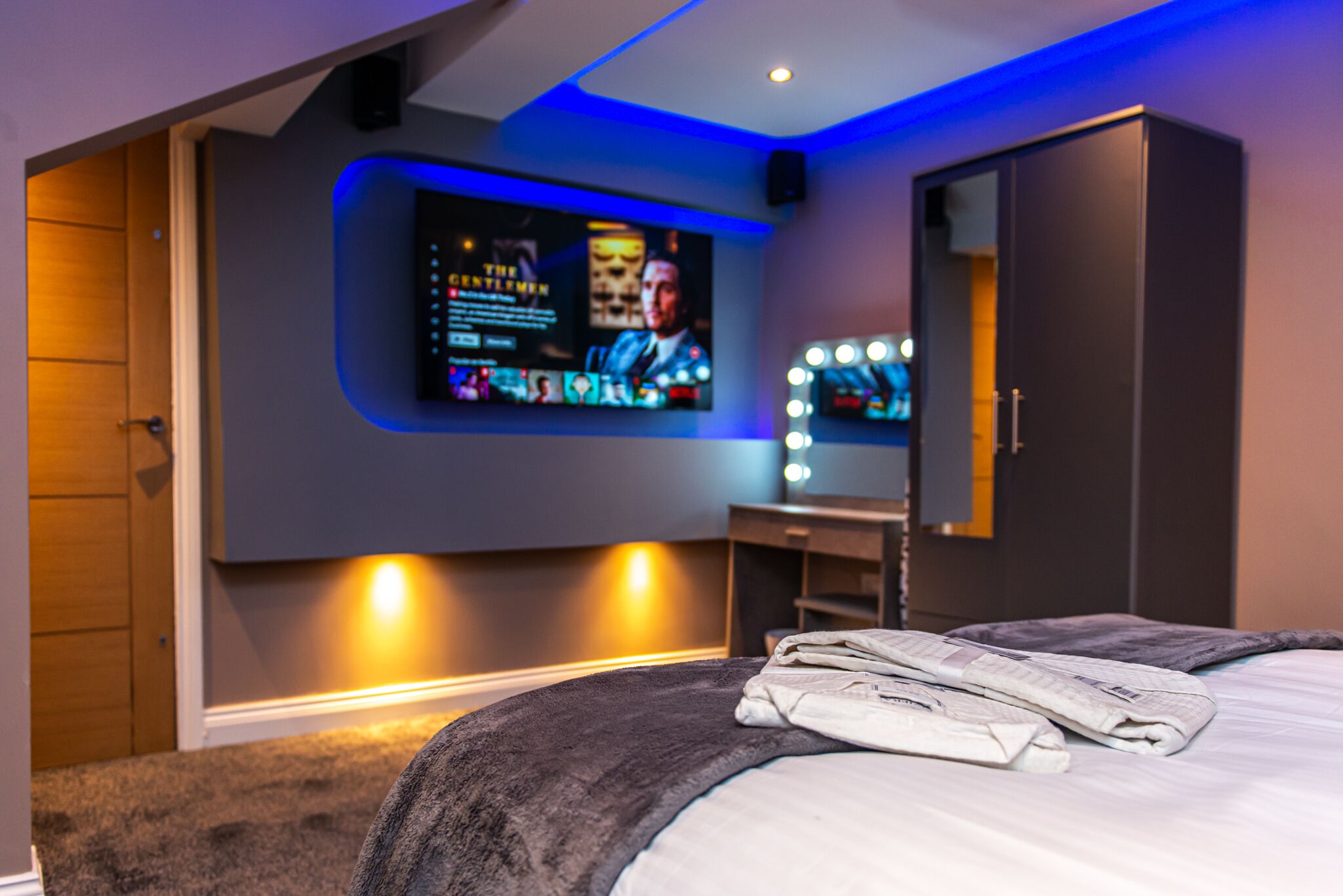 A modern bedroom with a large bed, a vanity with lights, and a wall-mounted TV displaying a movie.