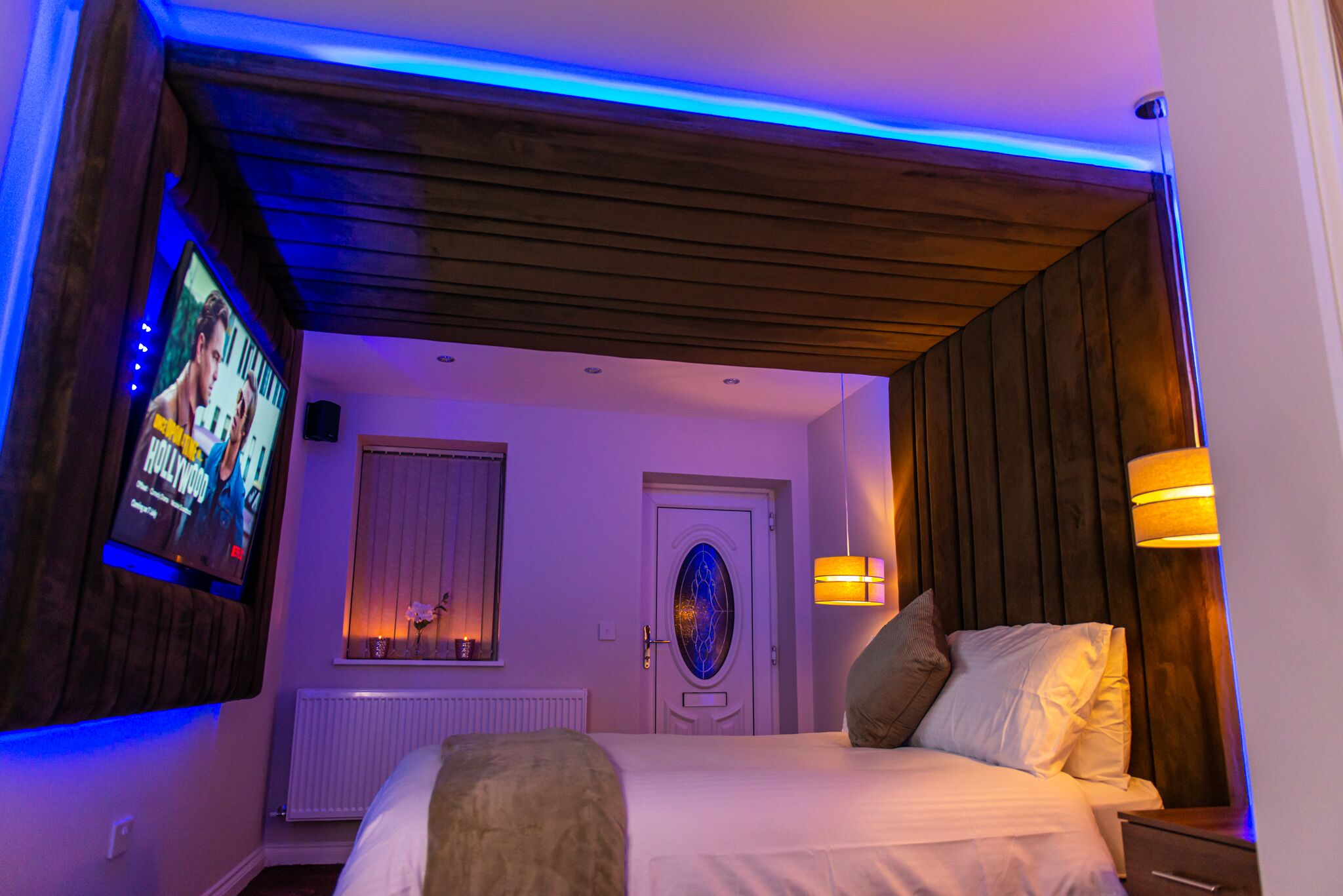 Habitación de hotel con una cama, un televisor en la pared, una puerta con panel de vidrio decorativo y una lámpara fijada a la pared.
