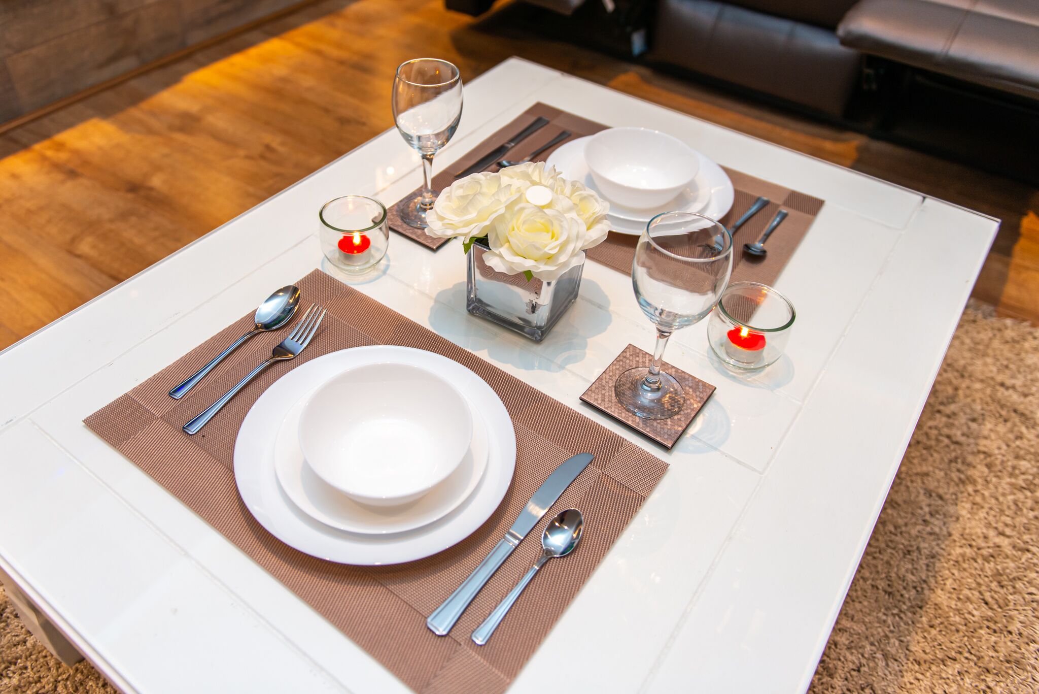 A neatly set dining table with white plates, silverware, and glasses, accompanied by a small vase of white flowers.