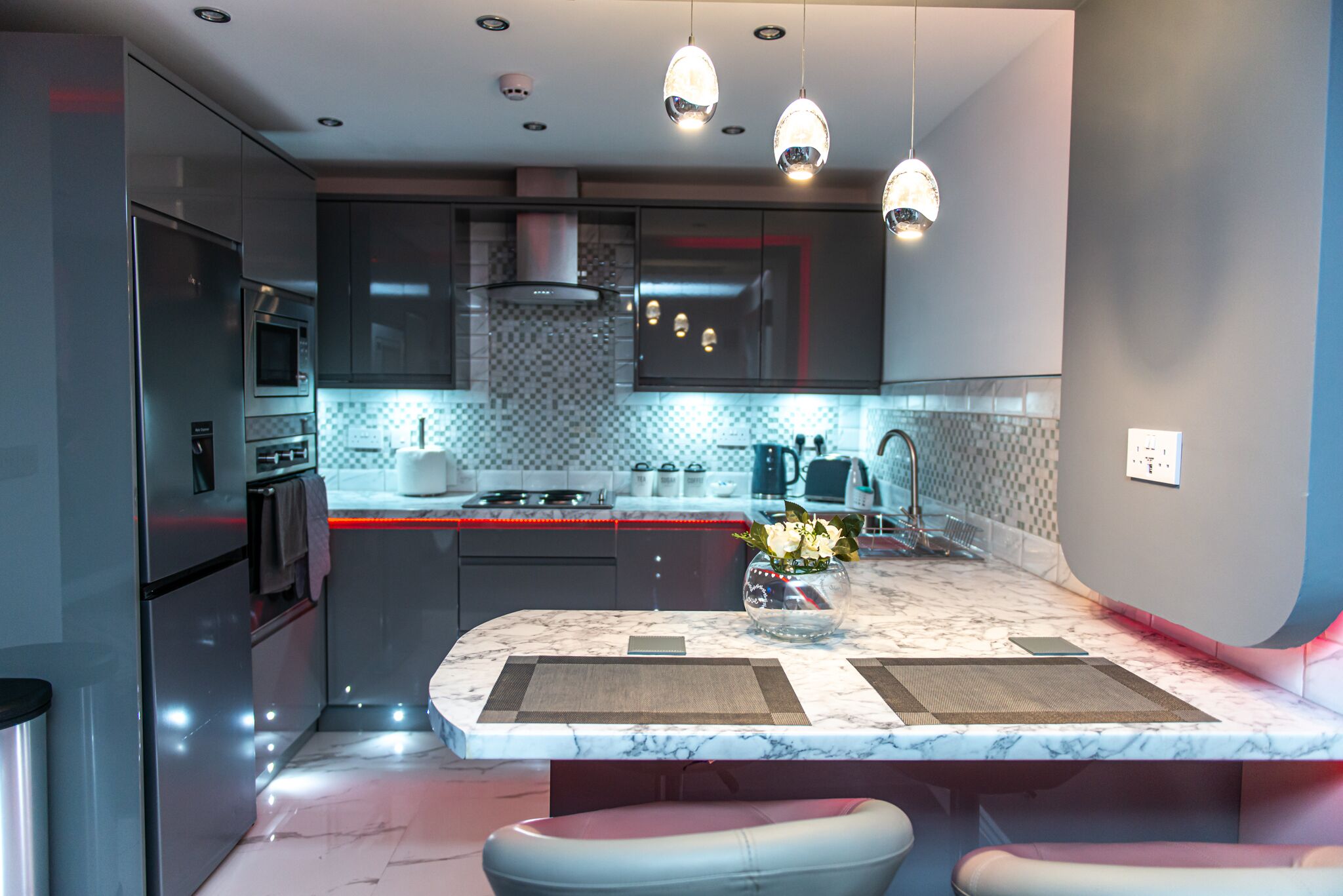 Una cocina moderna con encimera de mármol, electrodomésticos de acero inoxidable, iluminación colgante y una isla central con taburetes.