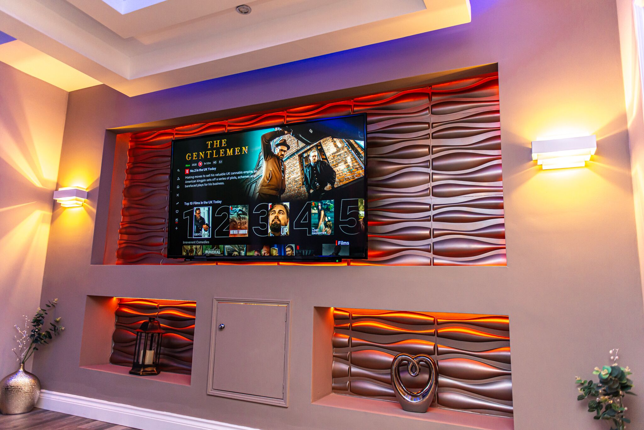 Pared con un televisor montado mostrando la película 'The Gentleman', paneles decorativos y dos lámparas de pared.