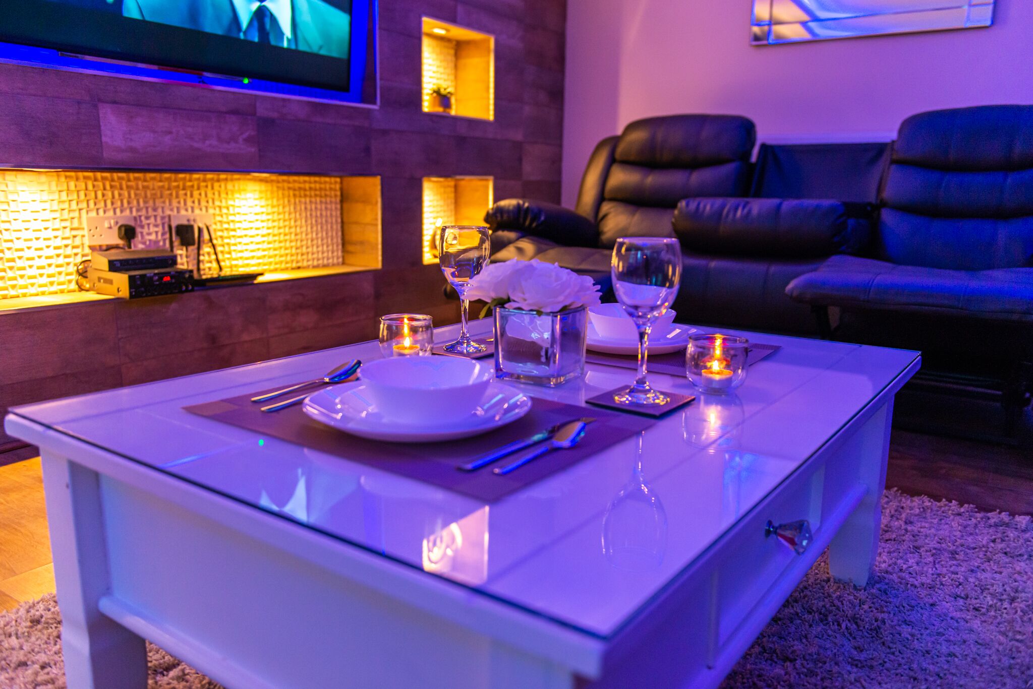A modern living room with a white coffee table set for dining, a leather sofa, and a flat-screen TV mounted on the wall.
