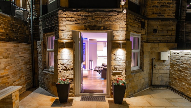 A stone-walled entrance with a bench, two potted plants, and a well-lit interior visible through the doorway.