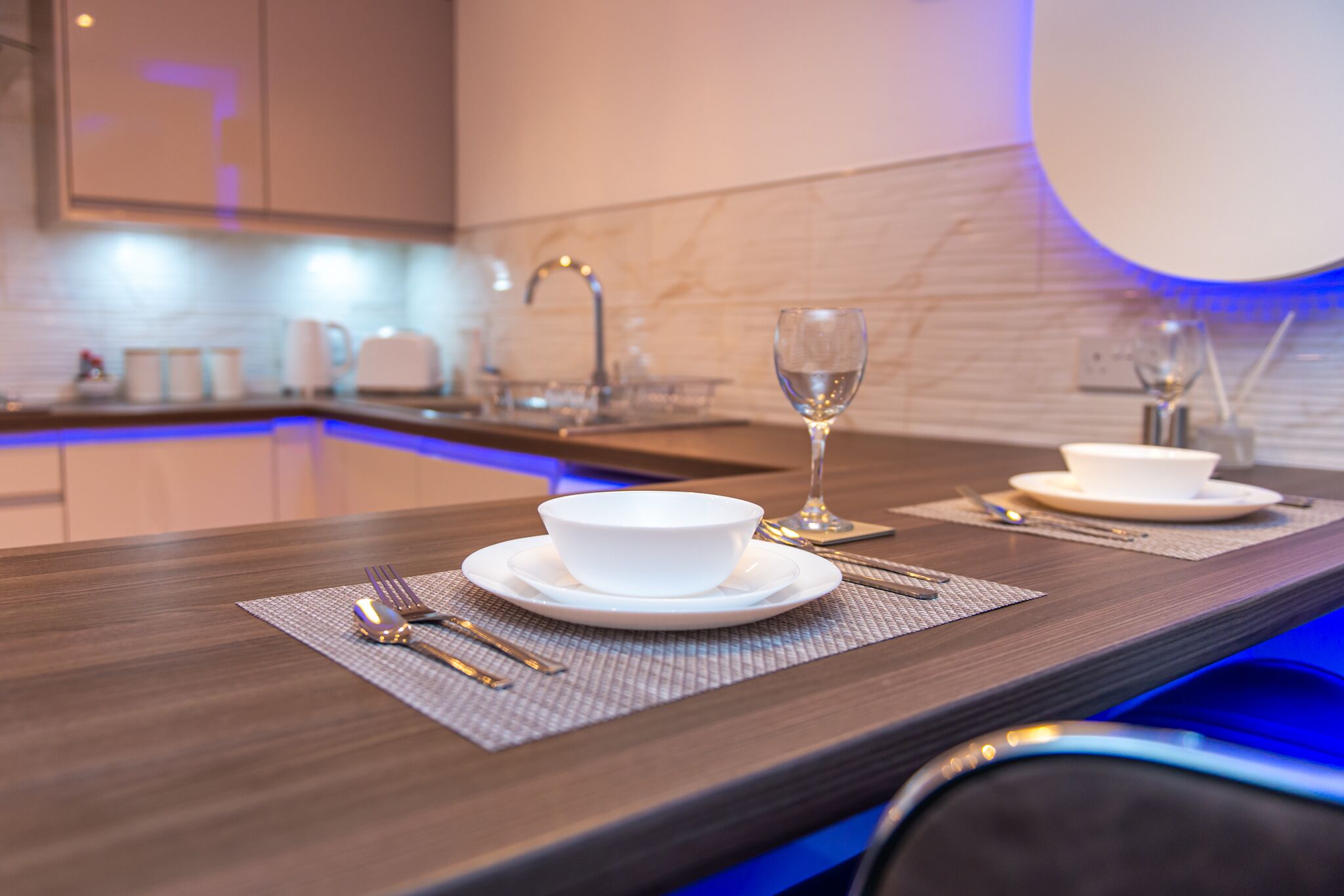 A modern kitchen with a dark countertop, white bowls, wine glasses, and cutlery on a placemat.