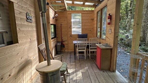 A wooden cabin with a central wood-burning stove, a dining area with a table and chairs, and a window overlooking a forested area. - “Maybelline”, 1959 Vintage Camper by the Creek (Barnardsville)
