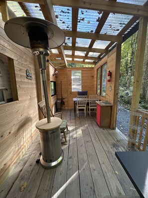 A wooden cabin with a central wood-burning stove, a dining area with a table and chairs, and a window overlooking a forested area. - “Maybelline”, 1959 Vintage Camper by the Creek (Barnardsville)
