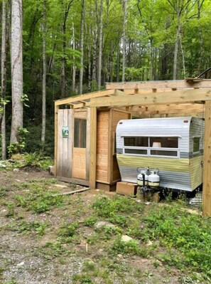 A small wooden cabin with a metal trailer attached, surrounded by a forest. - “Maybelline”, 1959 Vintage Camper by the Creek (Barnardsville)